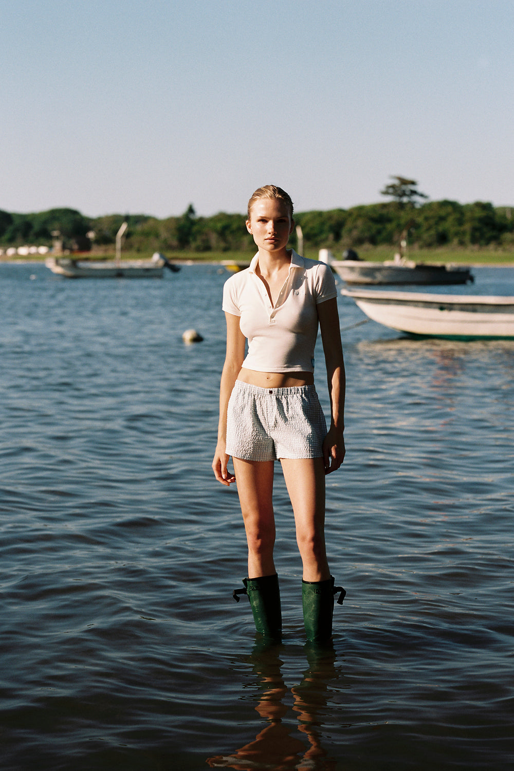 Woman standing in water wearing a white shirt, shorts, and green rain boots with boats and trees in the background.