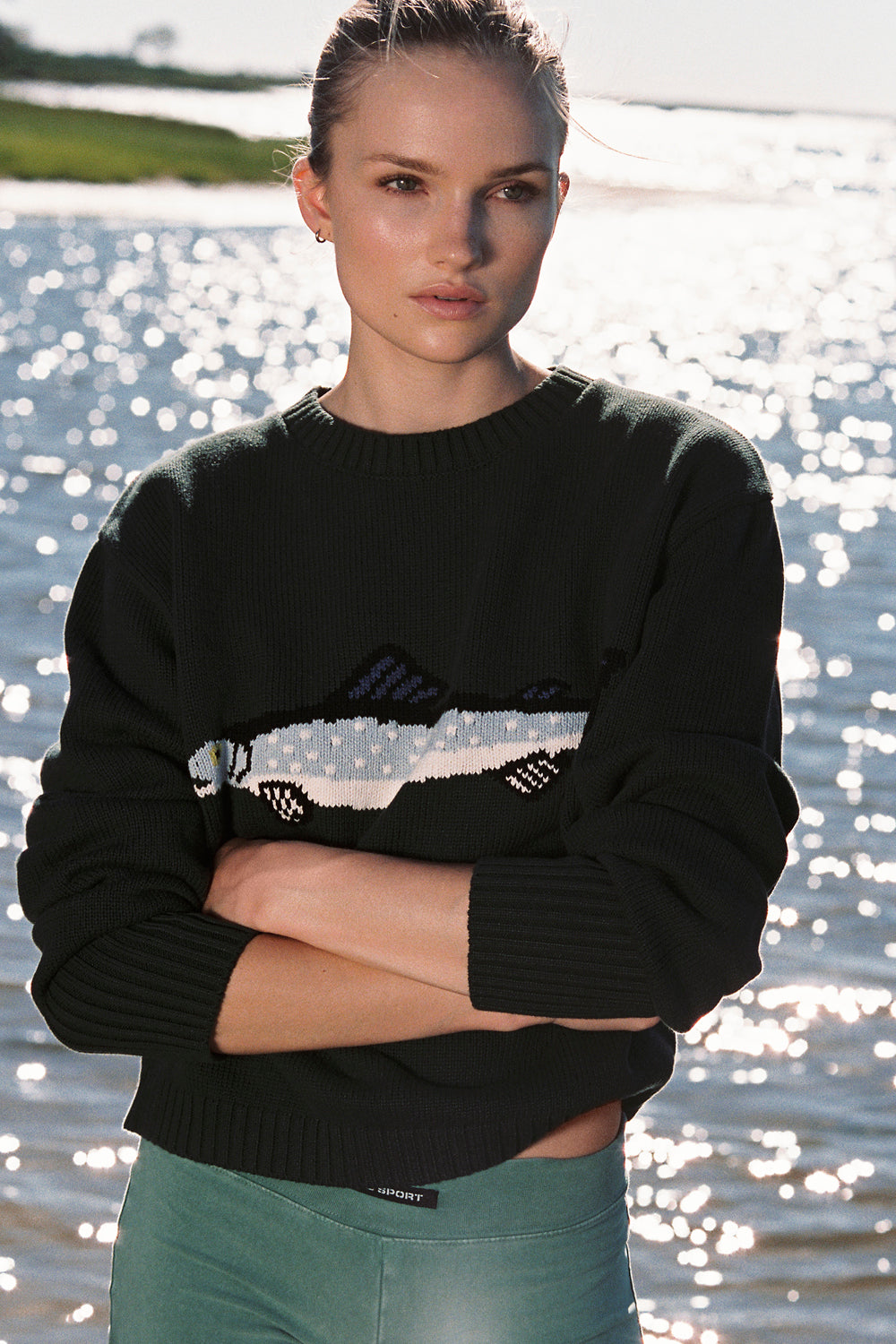 Woman wearing a black sweater with a fish design by a body of water.