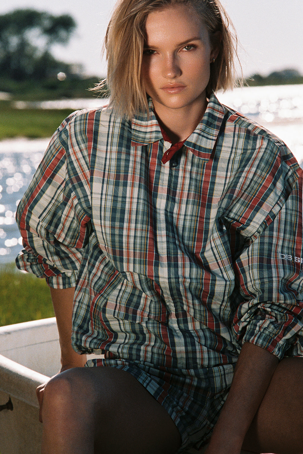 Woman wearing a plaid jacket sitting by a body of water.