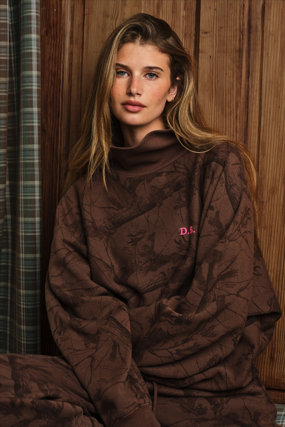 Model sits wearing brown camo rollneck sweatshirt with pink "D.B" monogram logo on the chest.
|M1