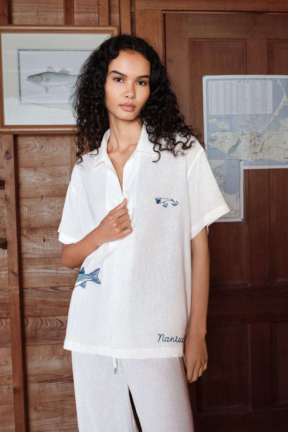 Woman wearing a white outfit with a brand logo in a room with wooden walls and framed pictures.
|M1