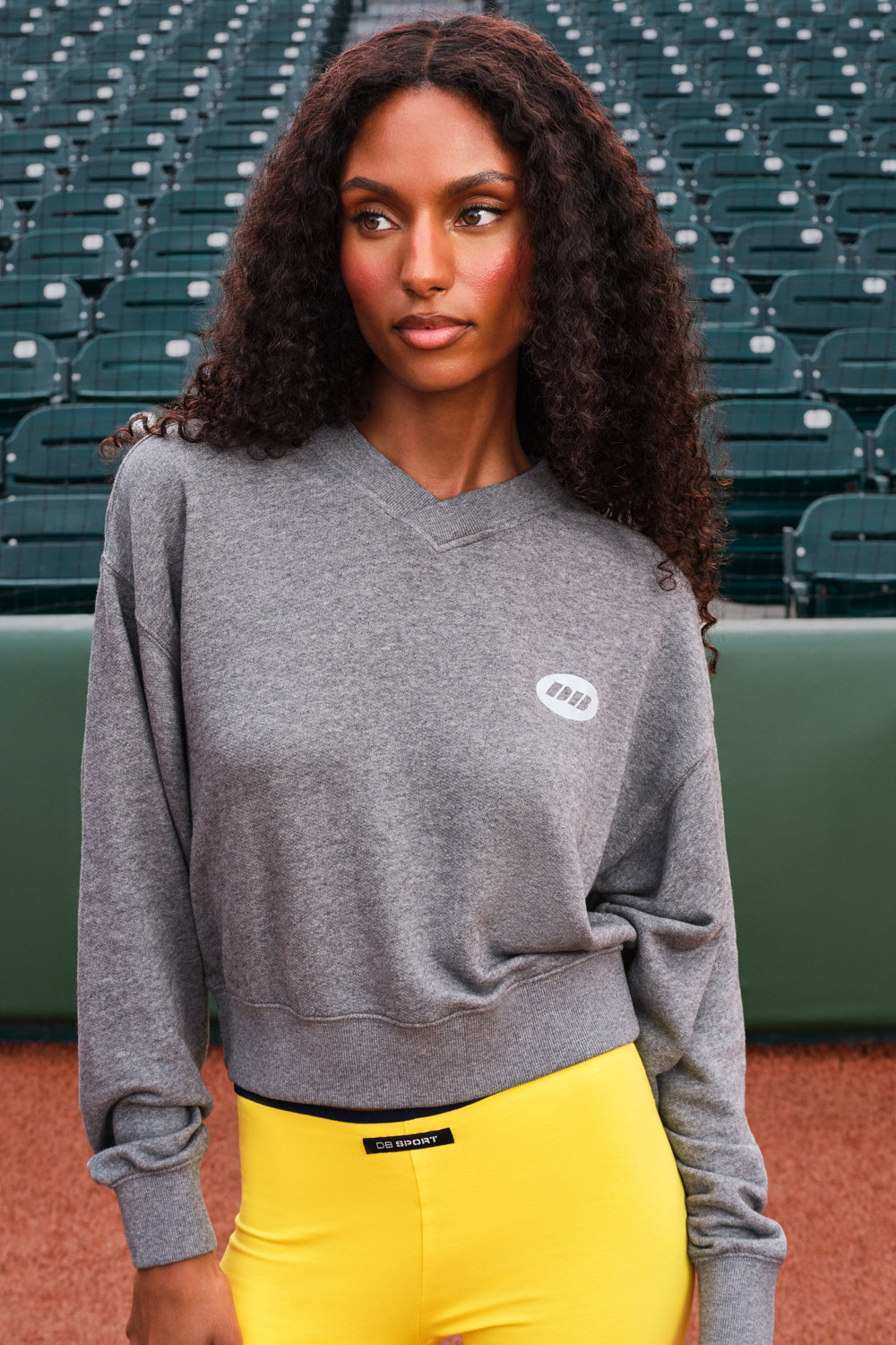 A woman with long curly dark hair stands in a stadium wearing a heather gray crewneck sweatshirt with a small white oval logo on the left chest, paired with bright yellow pants.
|M1