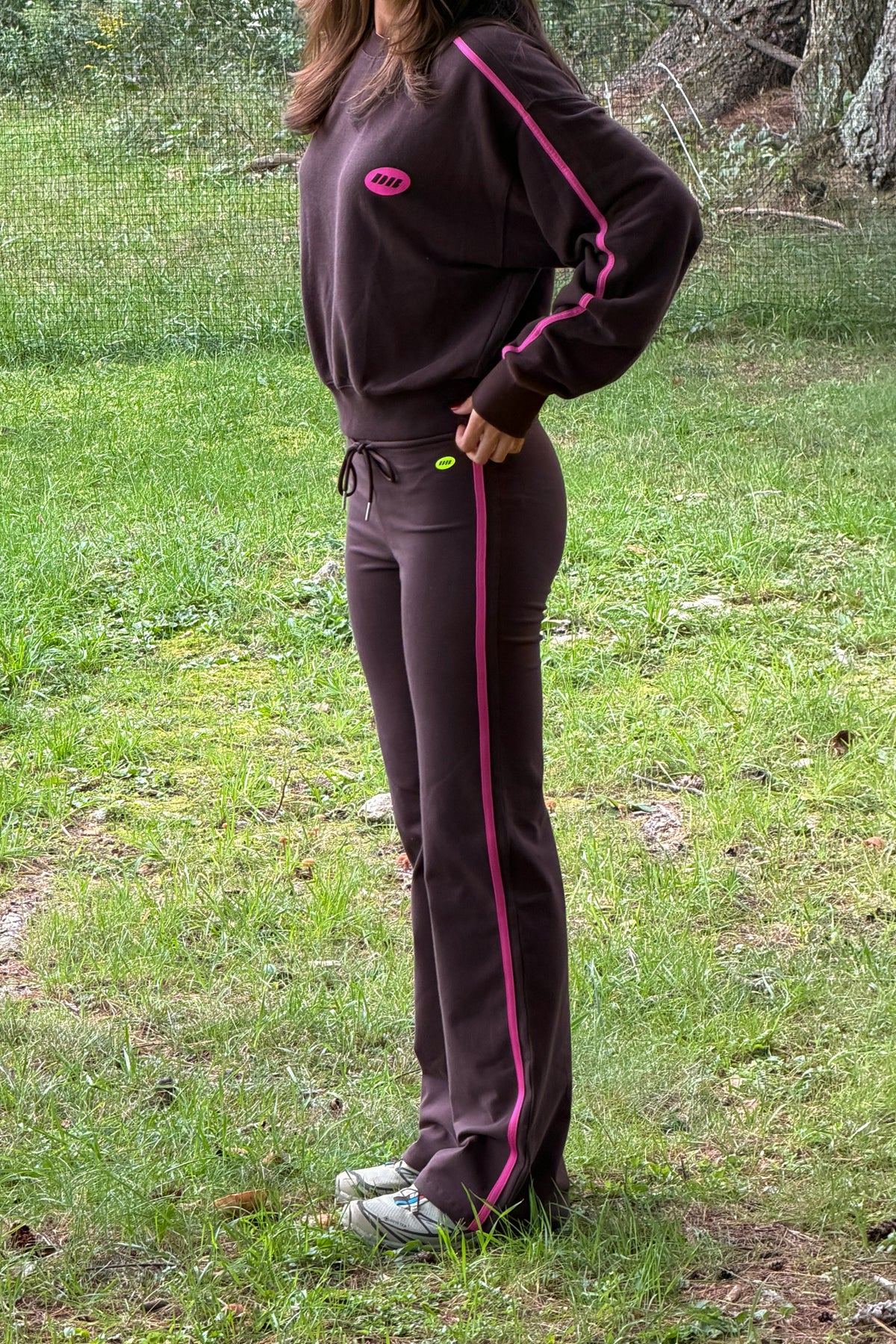 A person wearing a dark brown tracksuit with bright pink stripes and a pink chest logo, standing outdoors on a grassy lawn.
|M1