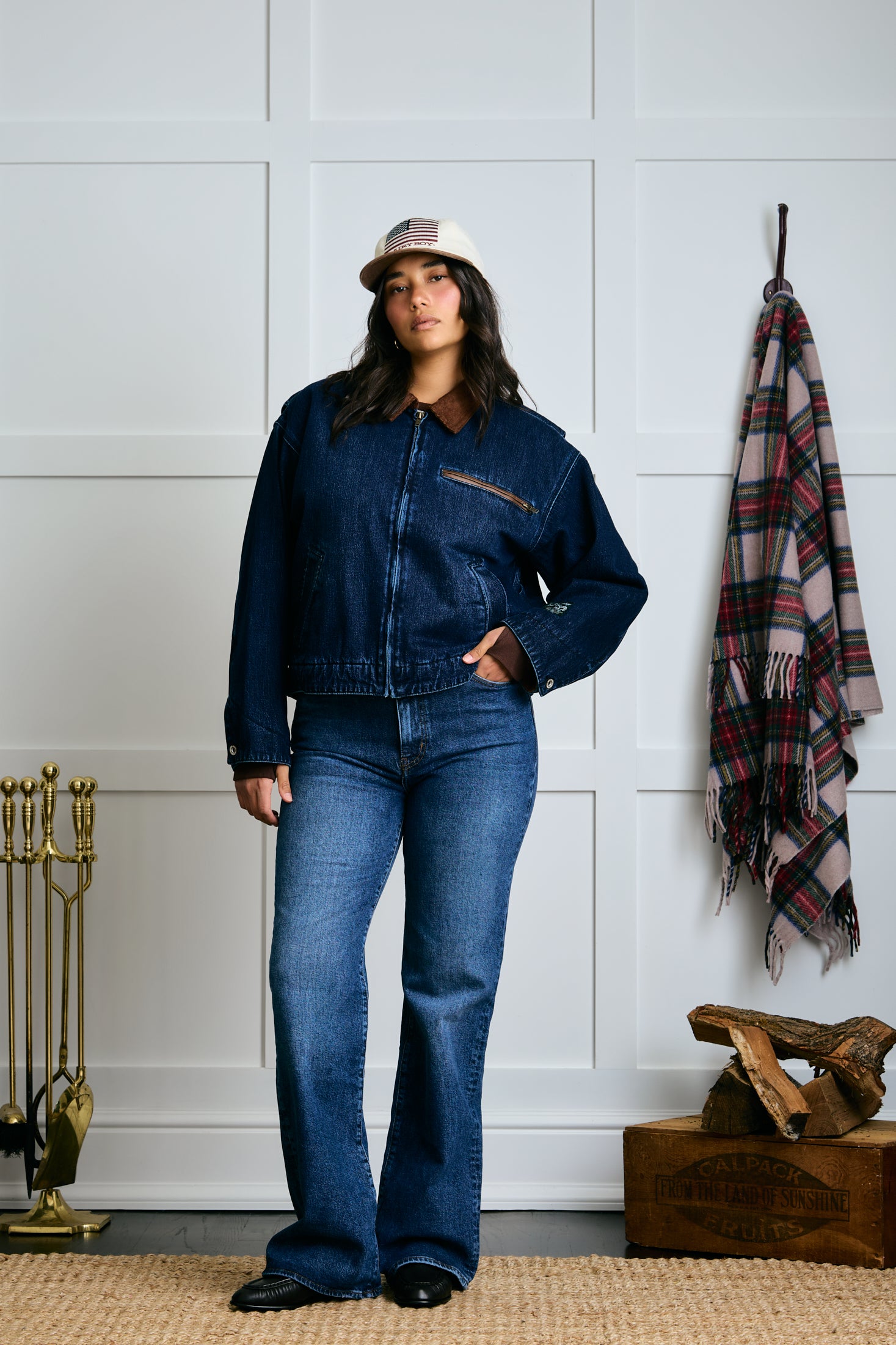 Woman wearing a denim jacket and jeans standing in a room with a plaid blanket and decorative items. |m1