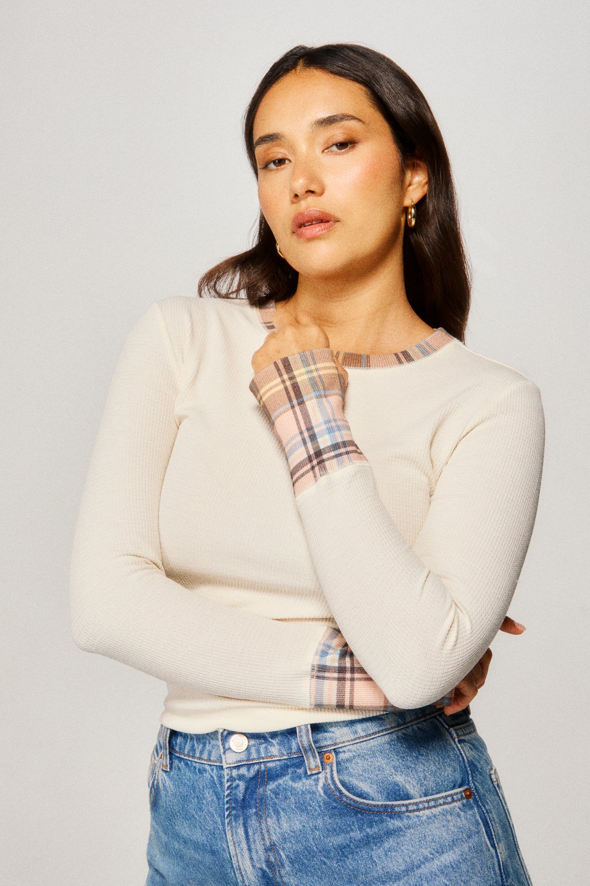 Woman wearing a cream sweater with plaid cuffs and blue jeans on a light gray background |m1