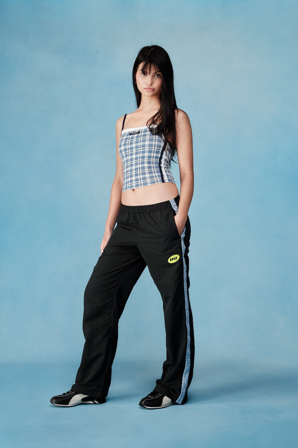 Woman wearing a plaid top and black pants with a logo on a blue background |M1