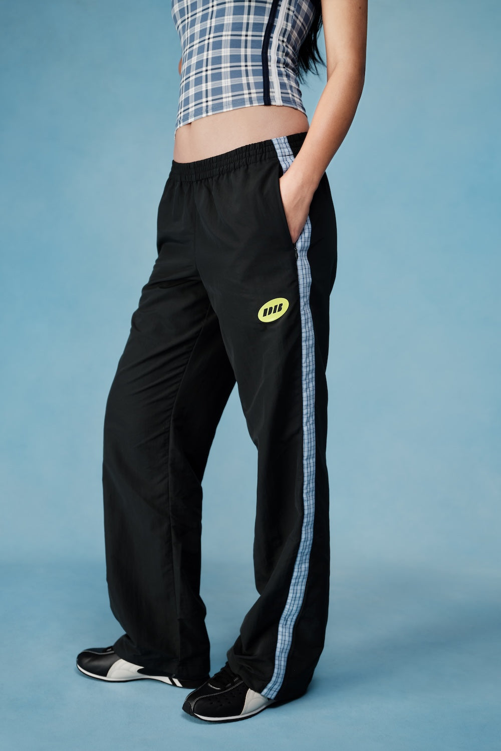 Person wearing black pants with a logo and a checkered top on a blue background |M1