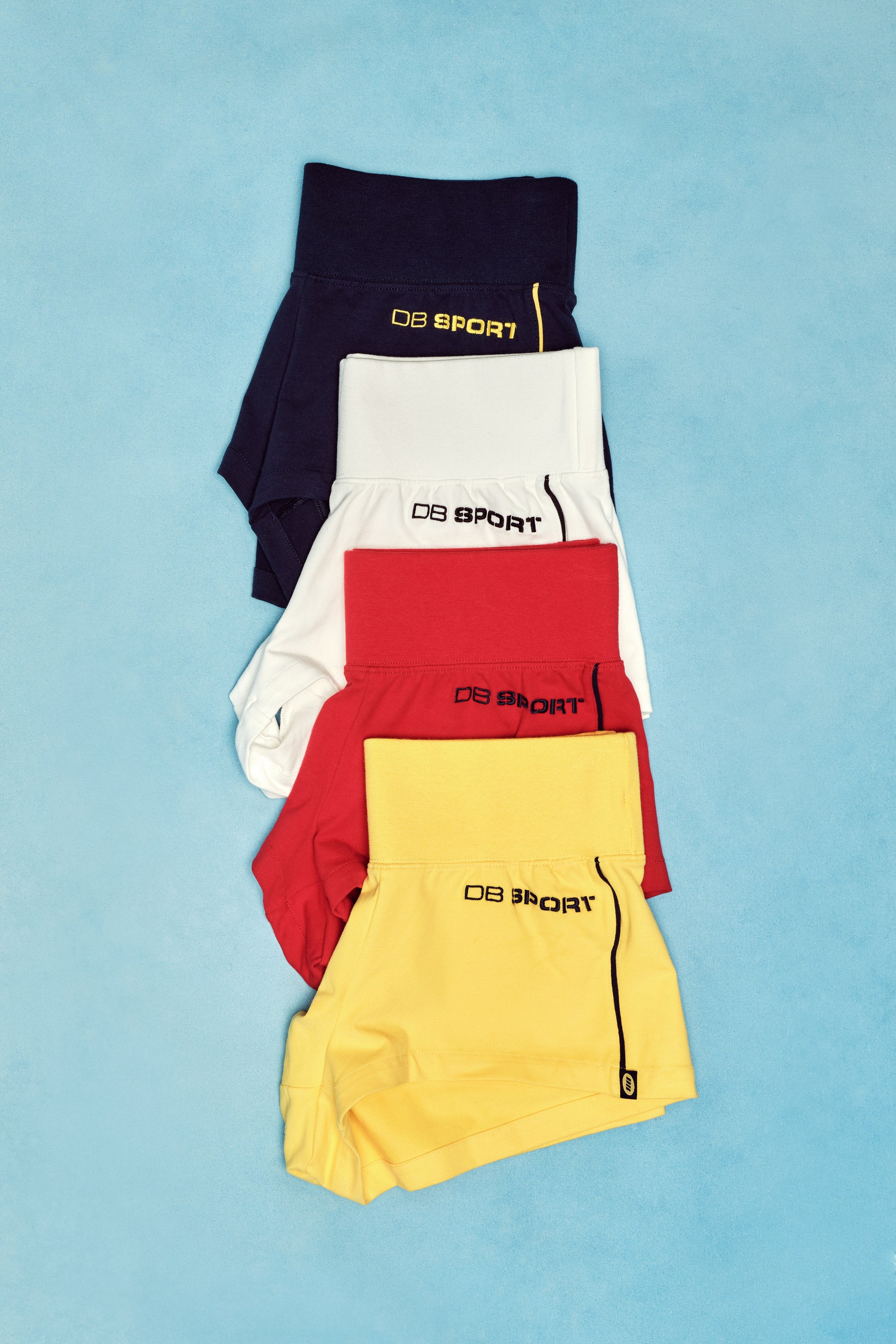 Four pairs of shorts in navy, white, red, and yellow with 'DB Sport' branding on a light blue background.