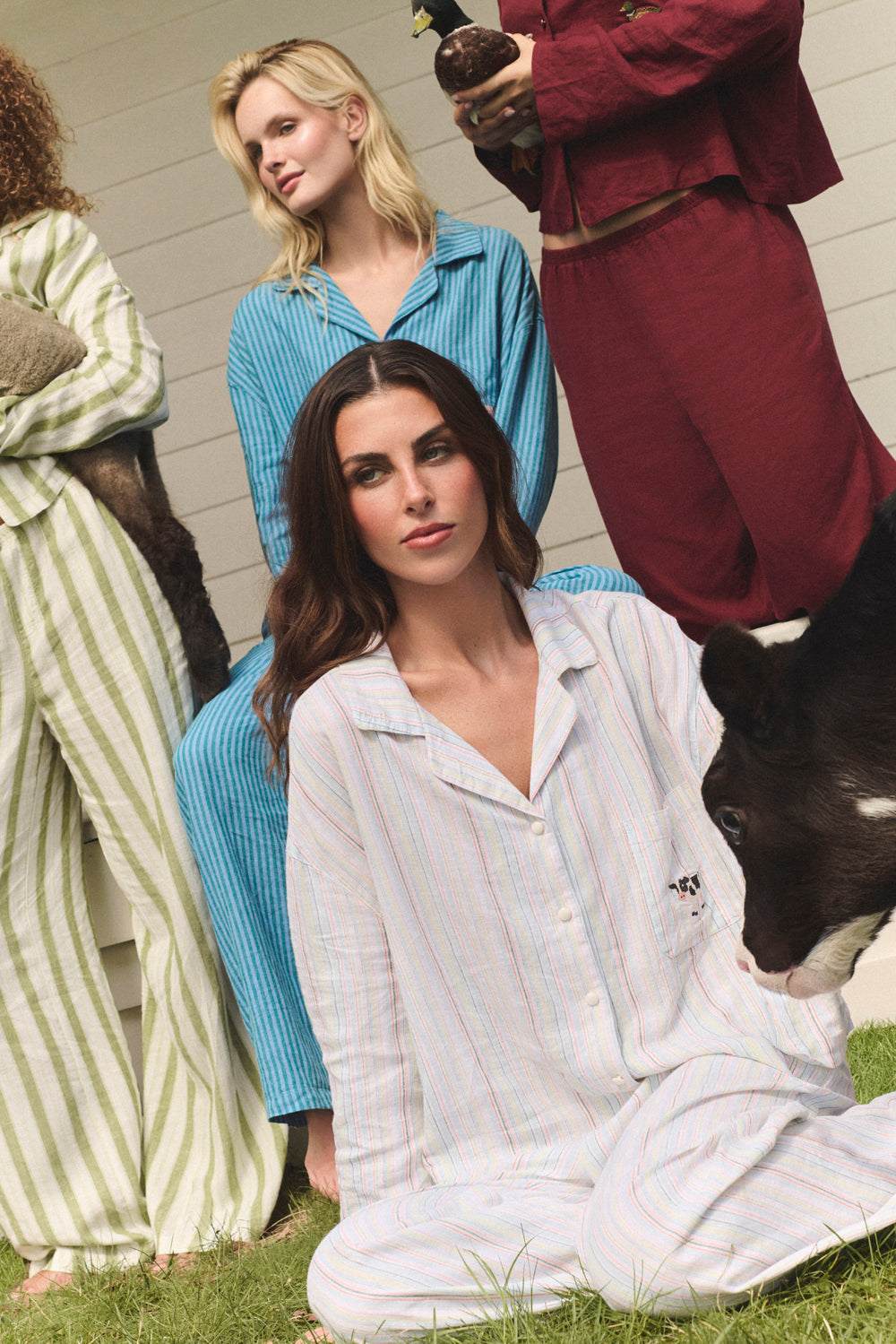 Woman in a striped pajama set sitting on grass with a cow, other people in colorful outfits in the background. |M1