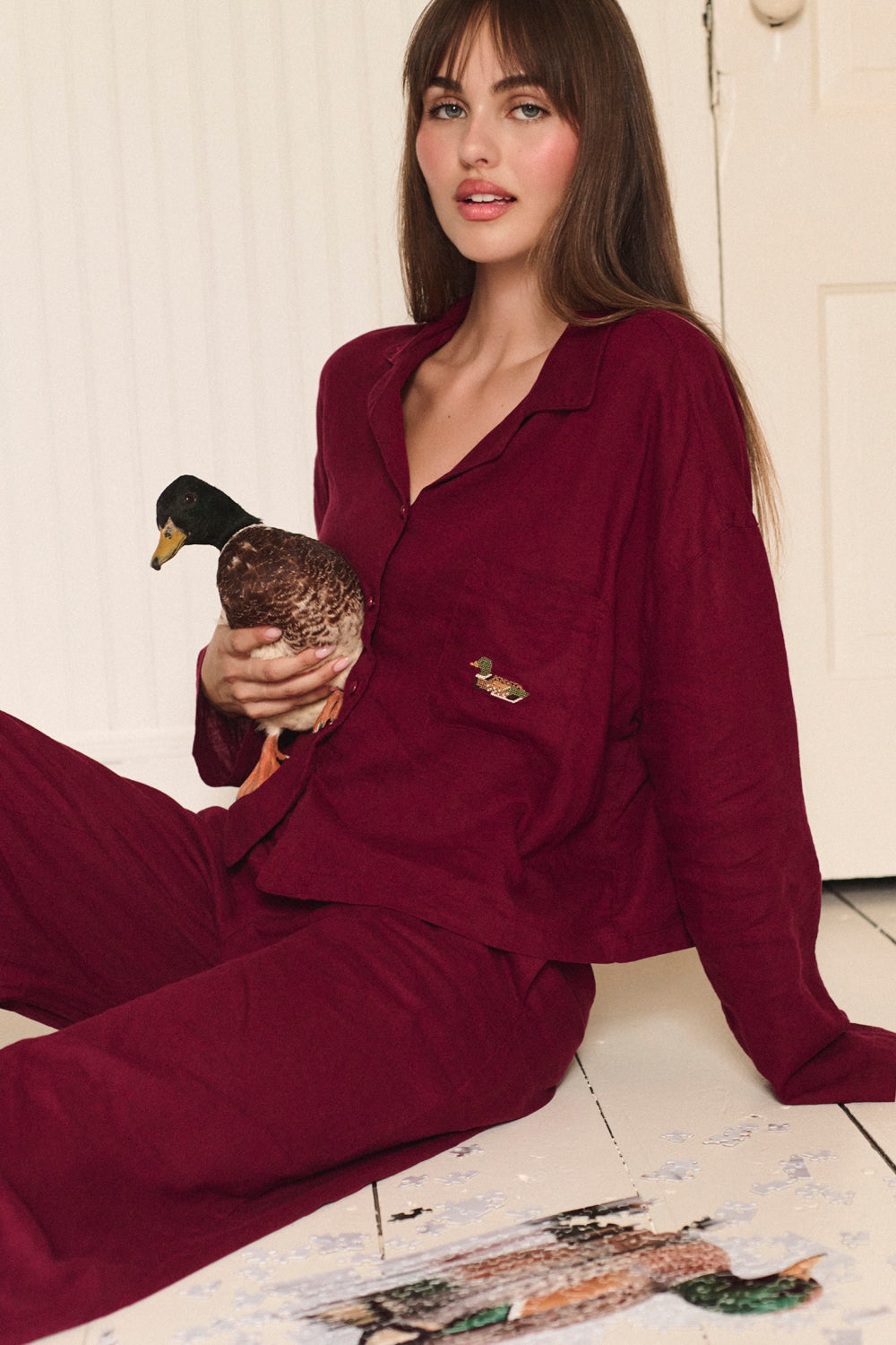 Woman in burgundy pajamas holding a duck on a wooden floor. |M1