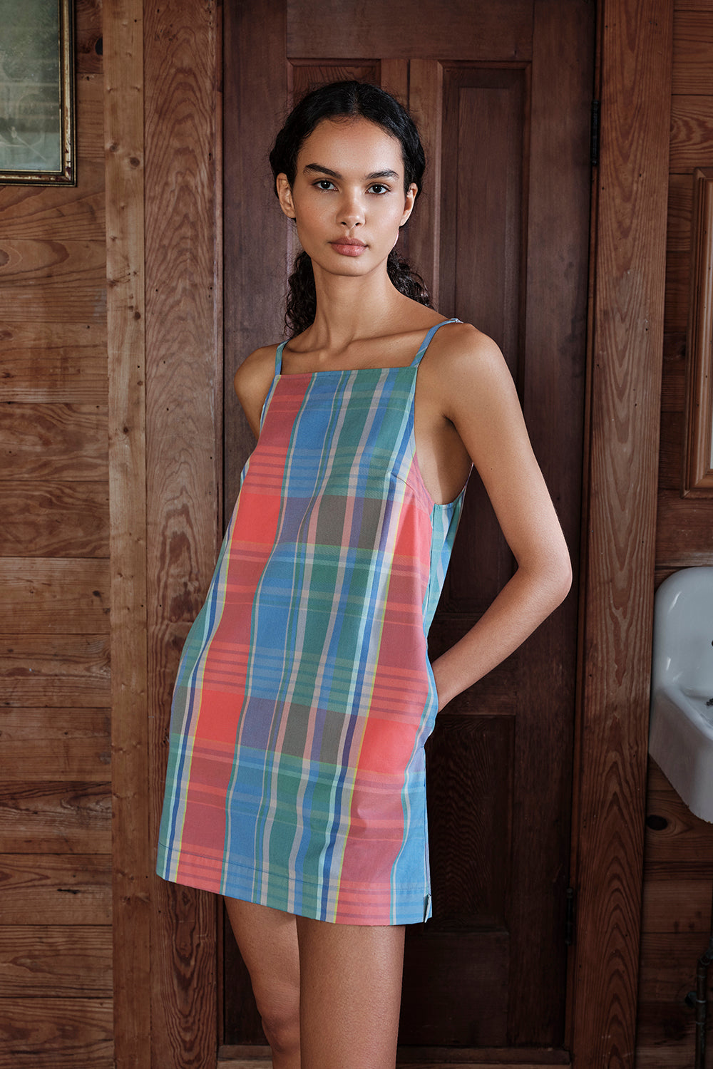 Woman wearing a colorful plaid dress standing in a wooden interior setting.
|M1