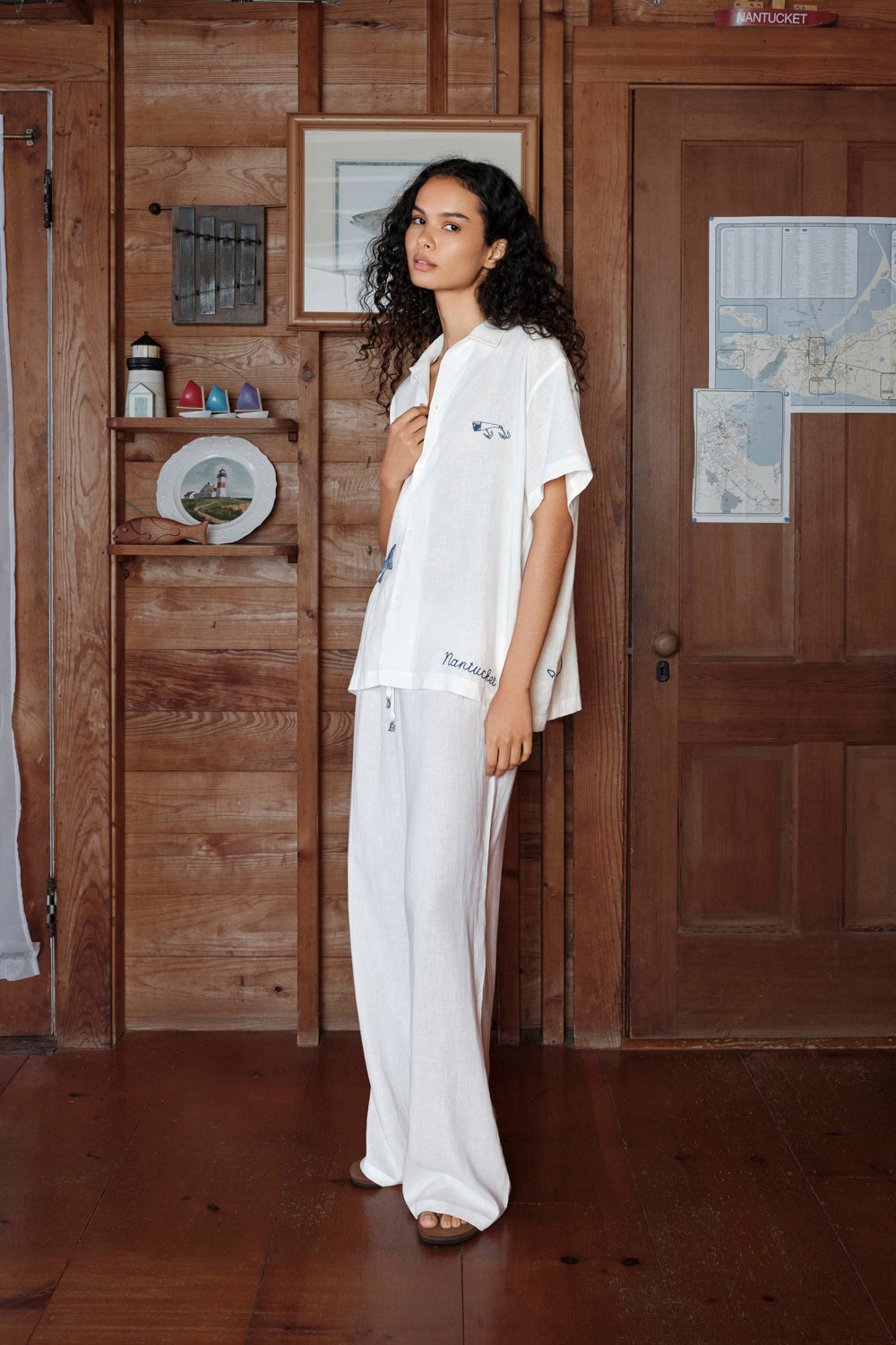 Woman in a white outfit standing in a room with wooden walls and a door.
|M1