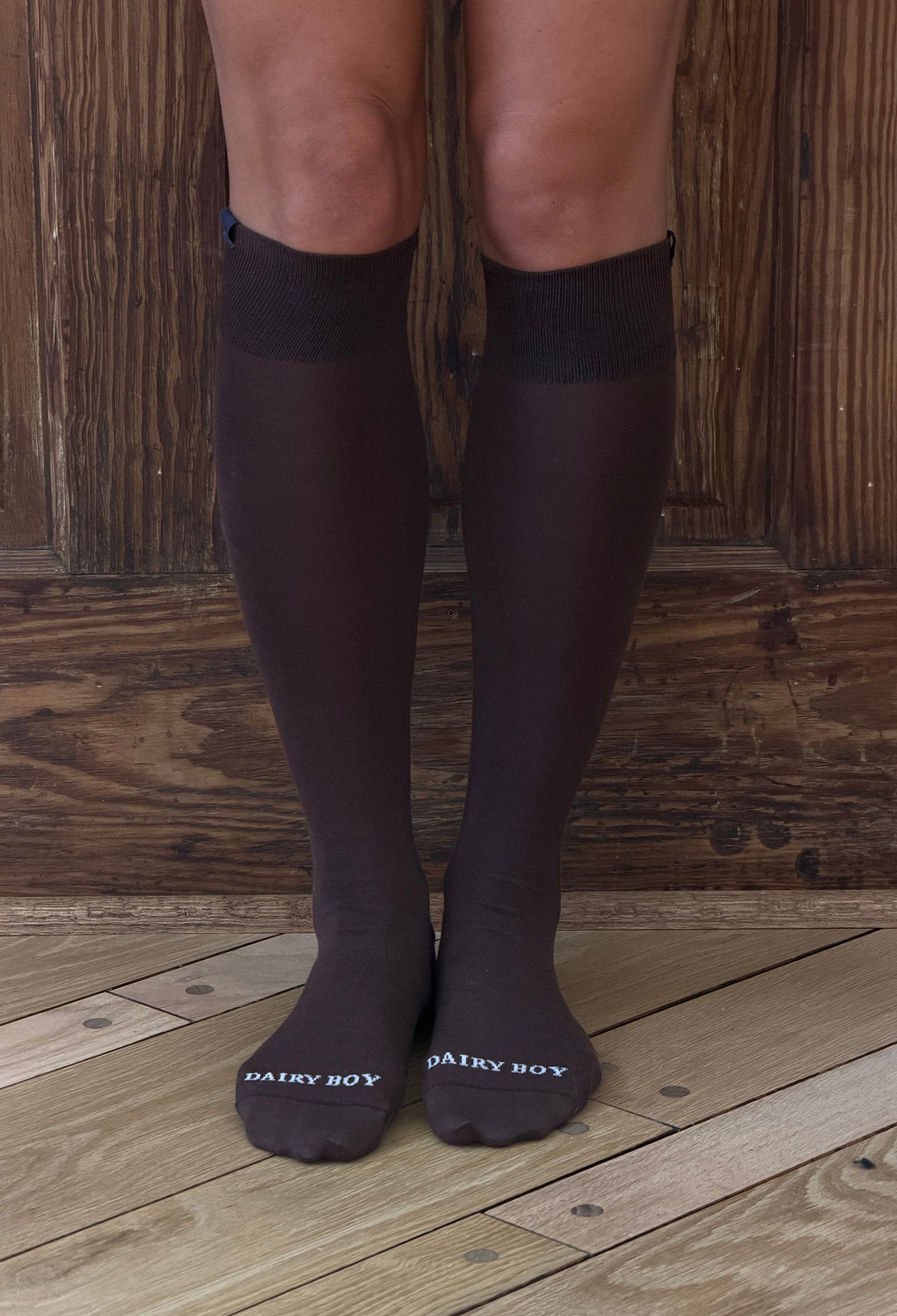 A person's legs wearing dark brown knee-high socks with white 'DAIRY BOY' branding on the toes, standing on a wooden plank floor in front of a rustic wooden wall.