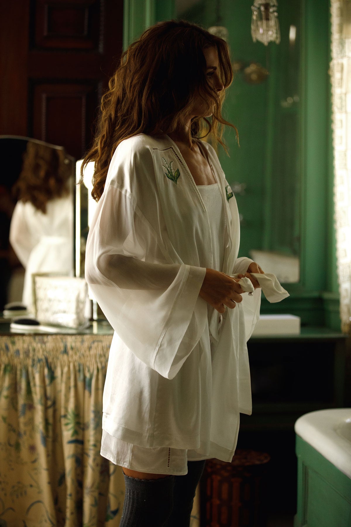 A woman in a white robe stands in a dimly lit bathroom or dressing area, holding a mirror while wearing dark thigh-high stockings.
|M1