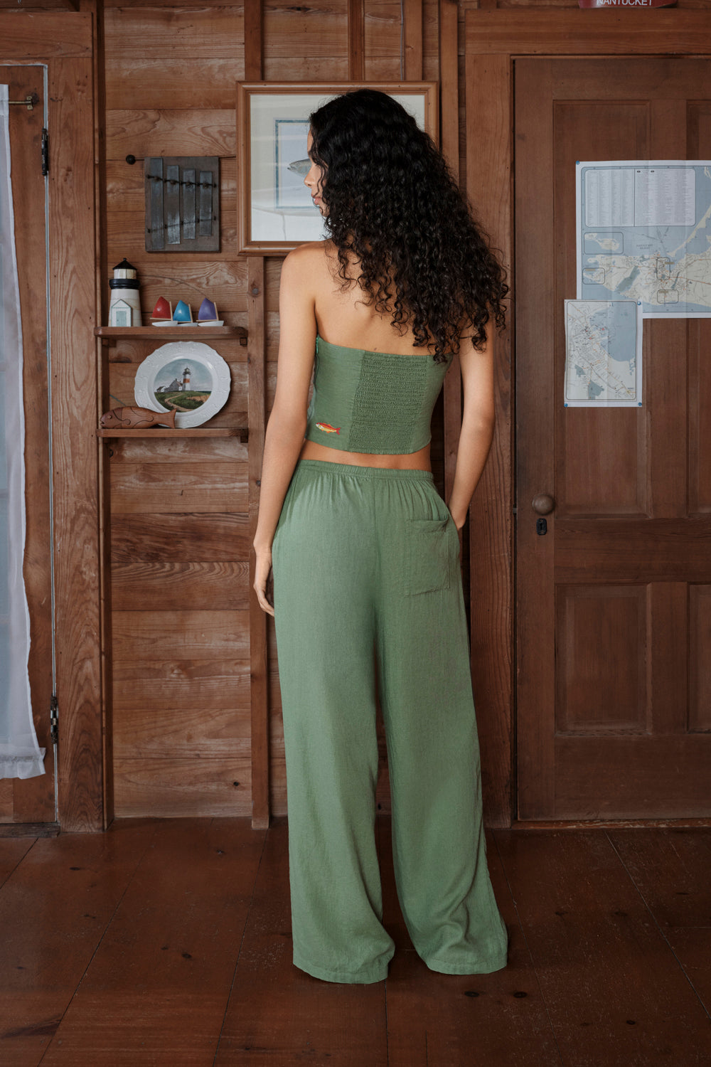 Woman in a green outfit standing in a room with wooden walls and a door.
|M1