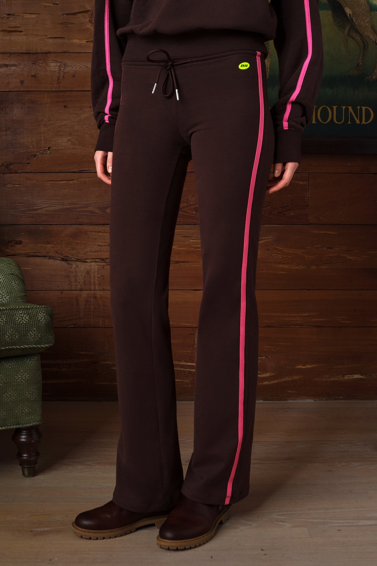 Dark brown wide-leg sweatpants with a vibrant pink stripe down the side and a matching pink stripe on the sleeve, shown on a model in front of a rustic wooden wall.
|M2