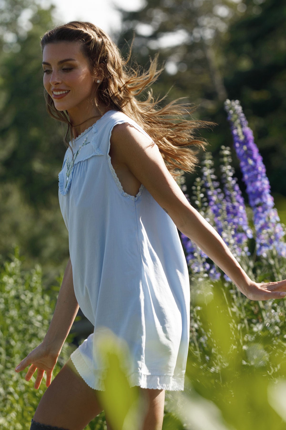 A woman wearing a light blue sleeveless tunic dress walking outdoors among green foliage and purple flowers. |M1