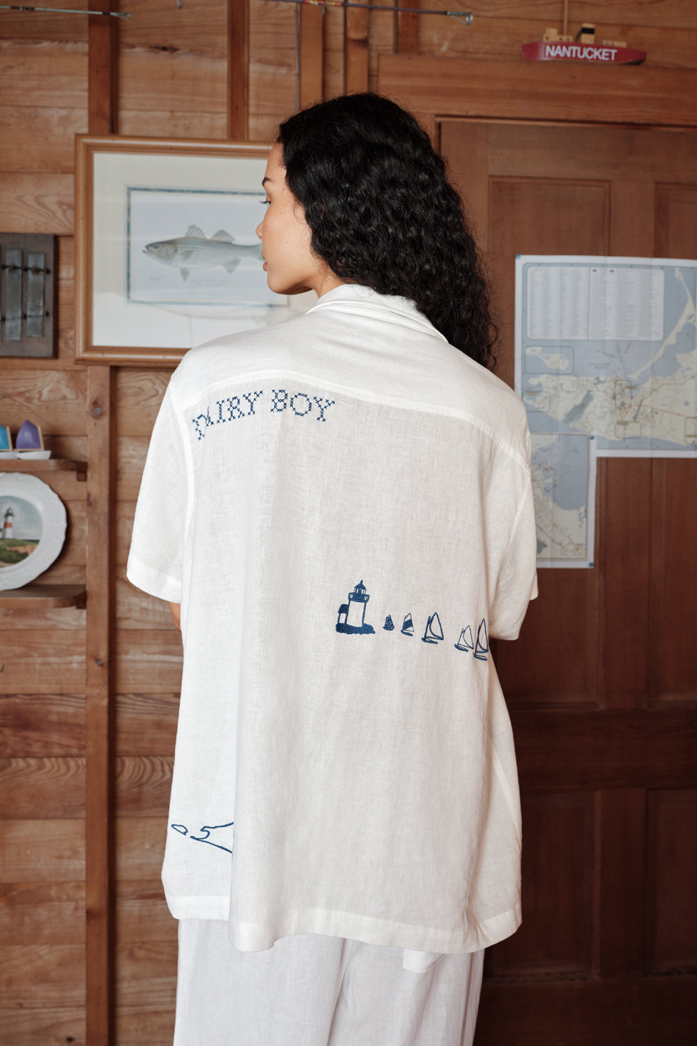 Person wearing a white shirt with embroidered designs in a room with wooden walls and decor.
|M1