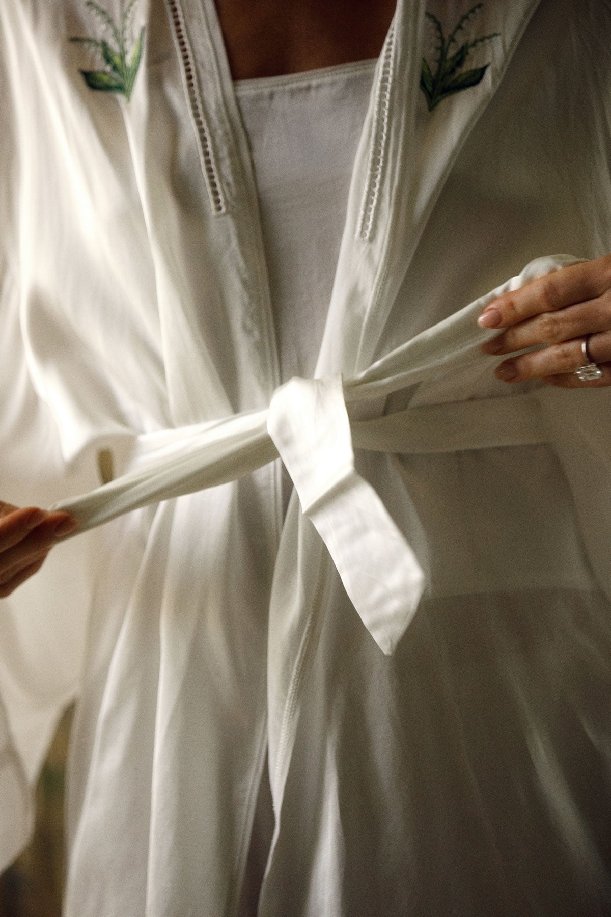 A close-up of a person wearing a white lightweight robe or kimono with embroidered green floral details near the neckline, tying the front tie with both hands.|M1