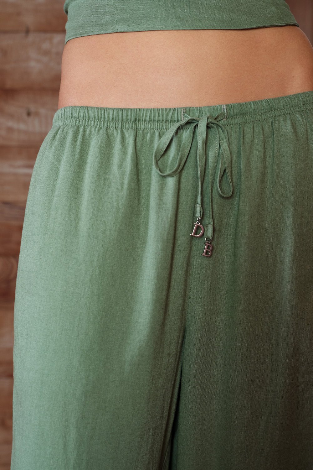 Green linen pants with a drawstring and brand logo on a wooden background
|M1