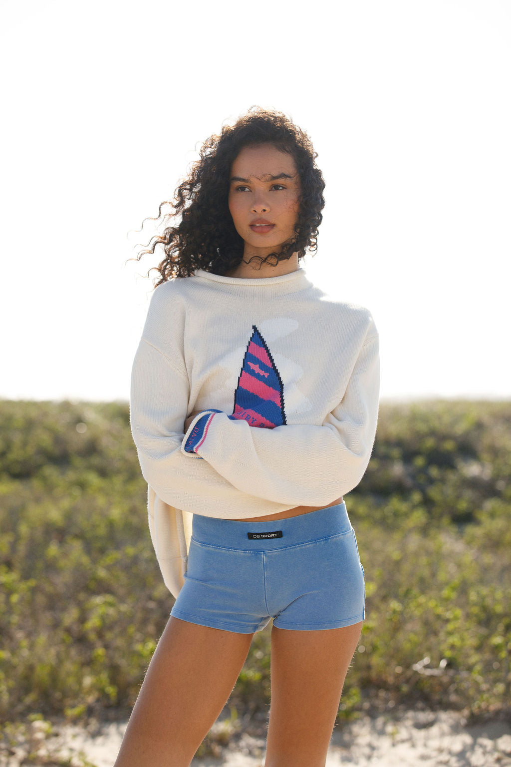 Woman wearing a white sweatshirt with a colorful design and blue shorts outdoors.
|M2