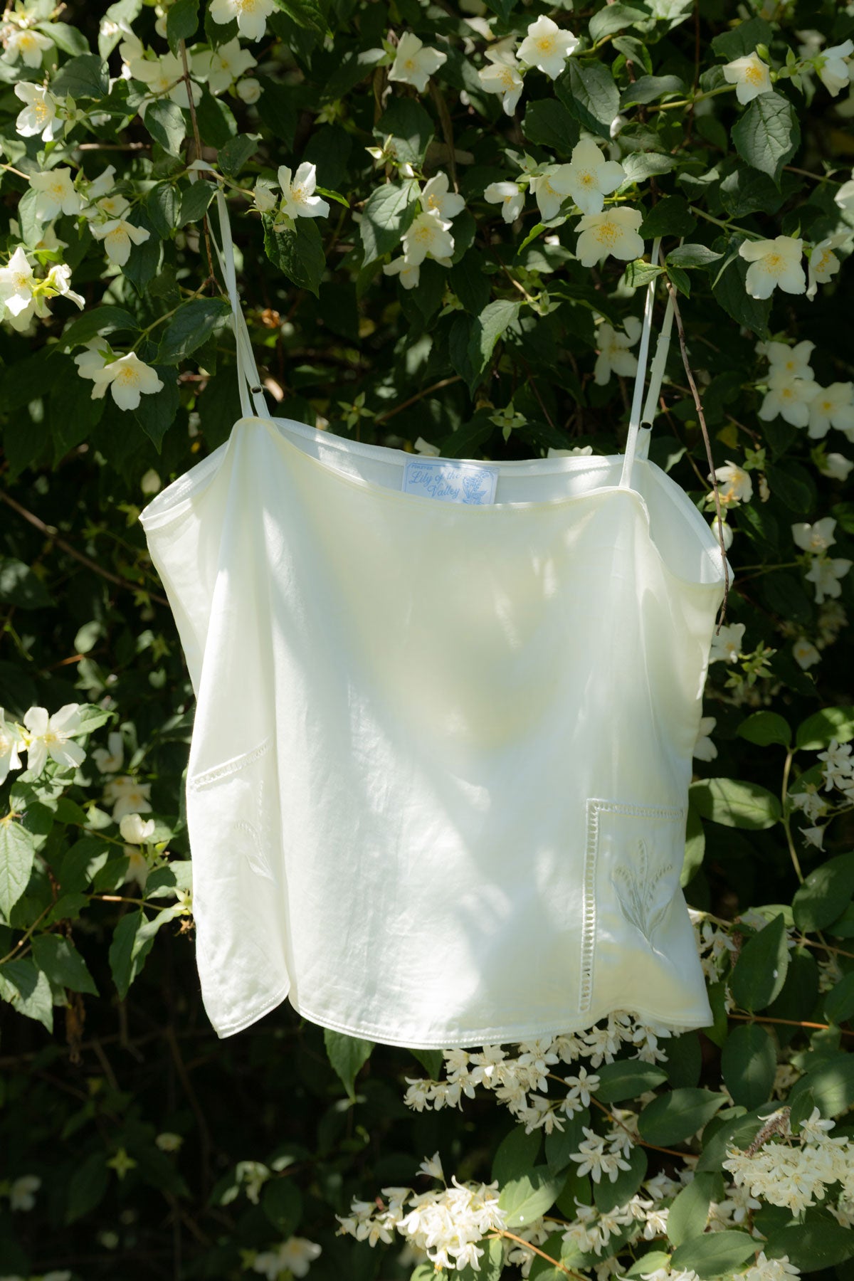 A white spaghetti-strap camisole top hanging outdoors against a background of green leaves and white flowers.