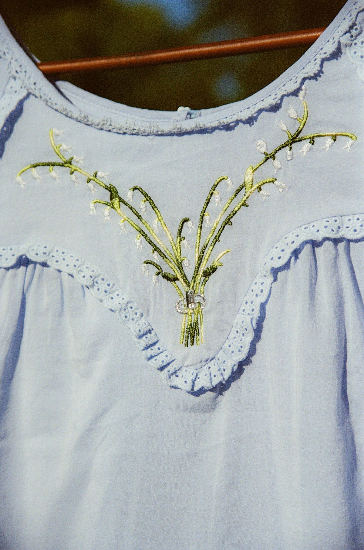 A close-up of a light blue embroidered blouse featuring a floral vine motif with green leaves and small white blossoms on the chest, finished with eyelet lace trim.