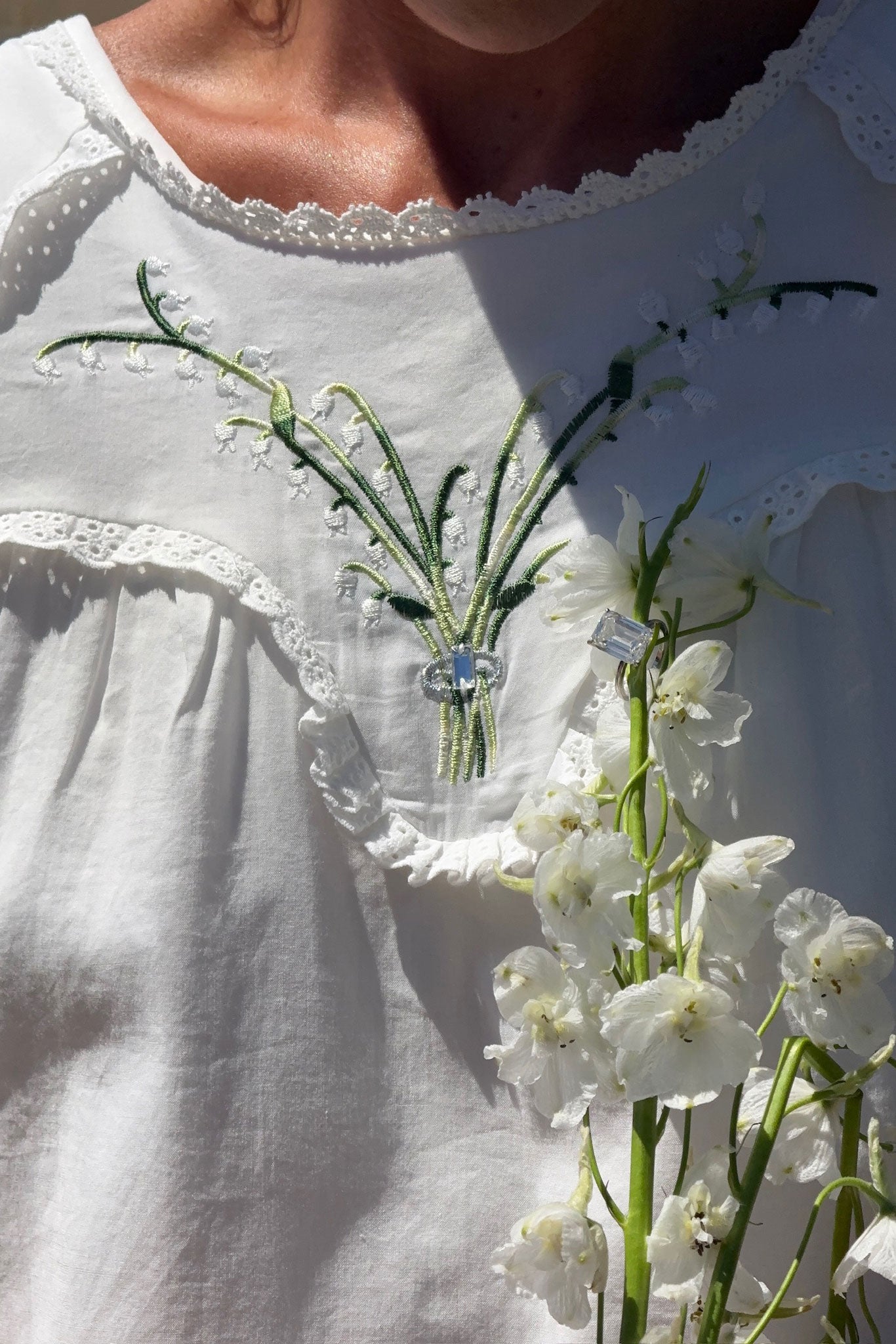 Close-up of a white embroidered dress with lace trim and green floral embroidery, worn by a model holding white flowers in bright sunlight.