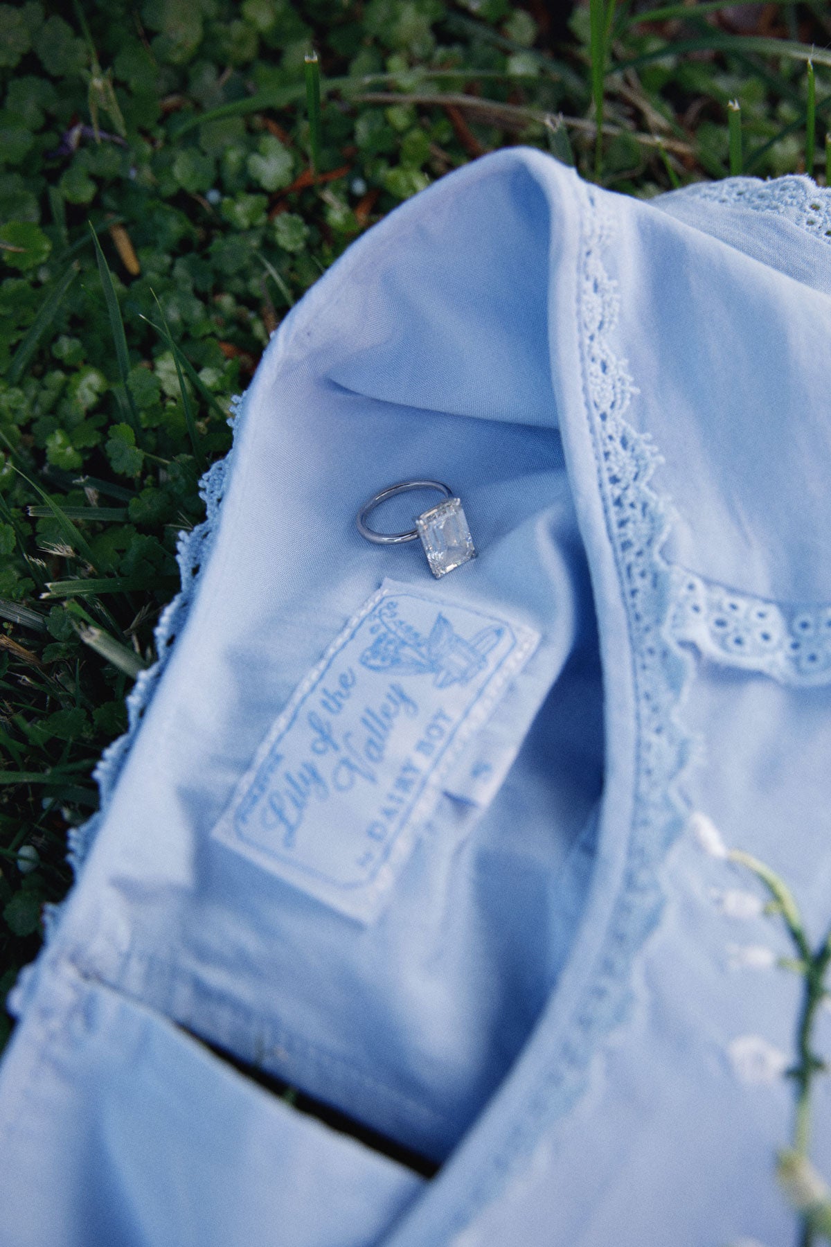 A light blue lace-trimmed garment laid on grass, with a small silver ring resting inside the opening. The inside label reads 'Lily of the Valley'