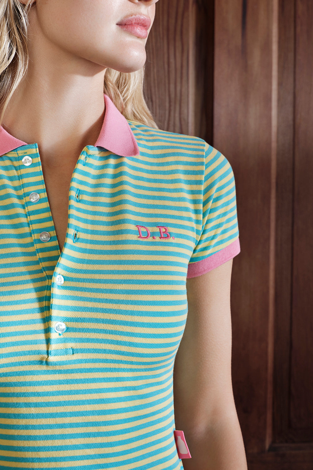 Woman wearing a striped polo shirt with pink collar and trim, featuring a logo on a wooden background.
|M1