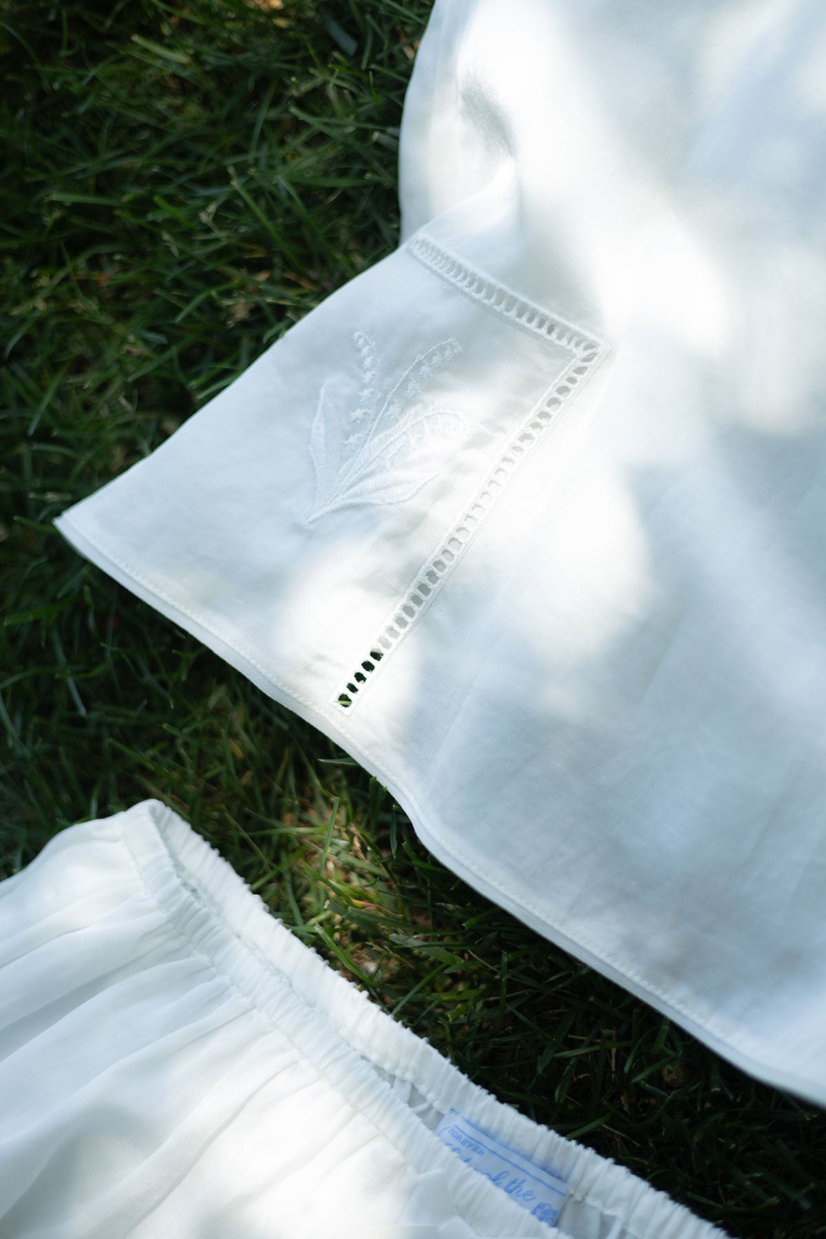 White embroidered fabric garment laid outdoors on green grass, showing a close-up of a sleeve or cuff with decorative stitching and a small embroidered motif.