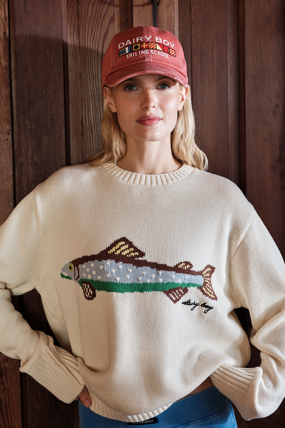 Woman wearing a cream sweater with a fish design and a red cap indoors.
|M1