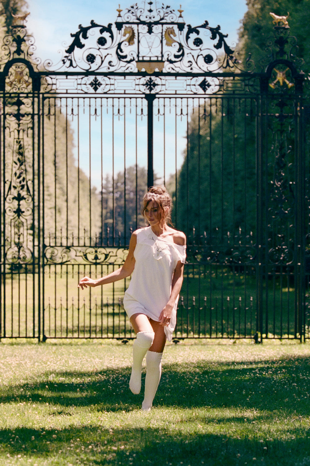 A woman in a white sleeveless dress and white thigh-high socks runs joyfully in front of an ornate black wrought-iron garden gate on a sunny day.
|M1
