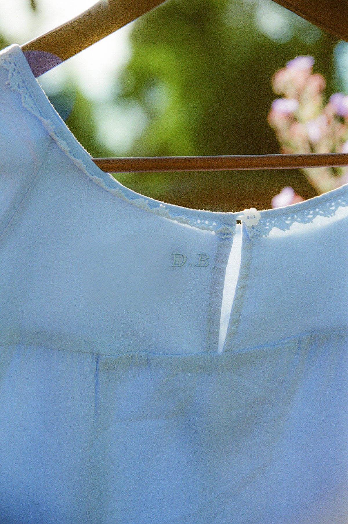 A light blue baby bodysuit or nightgown hanging on a wooden hanger against a blurred outdoor background with green foliage and pink flowers. The neckline features delicate lace trim and a small white button closure.