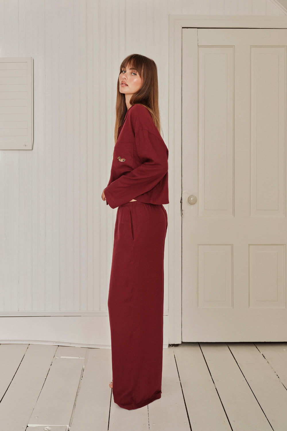 Woman wearing a burgundy pajama set standing in a room with white walls and a door. |M1
