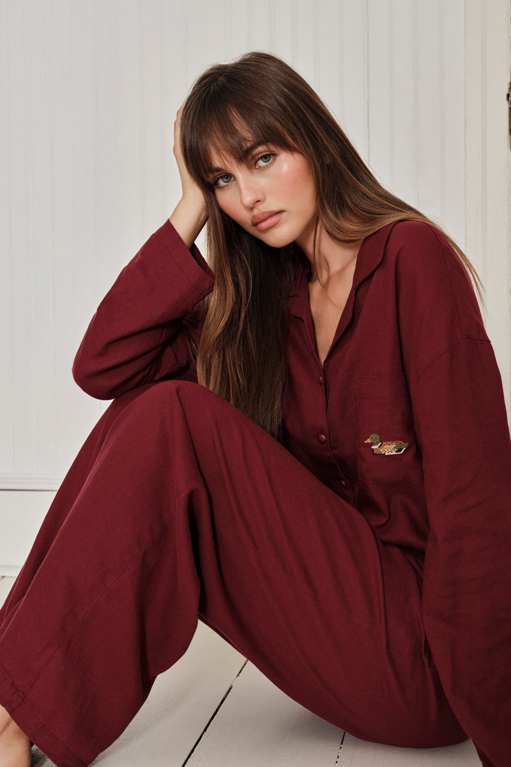 Woman wearing a burgundy pajama set sitting on a light wooden floor. |M1