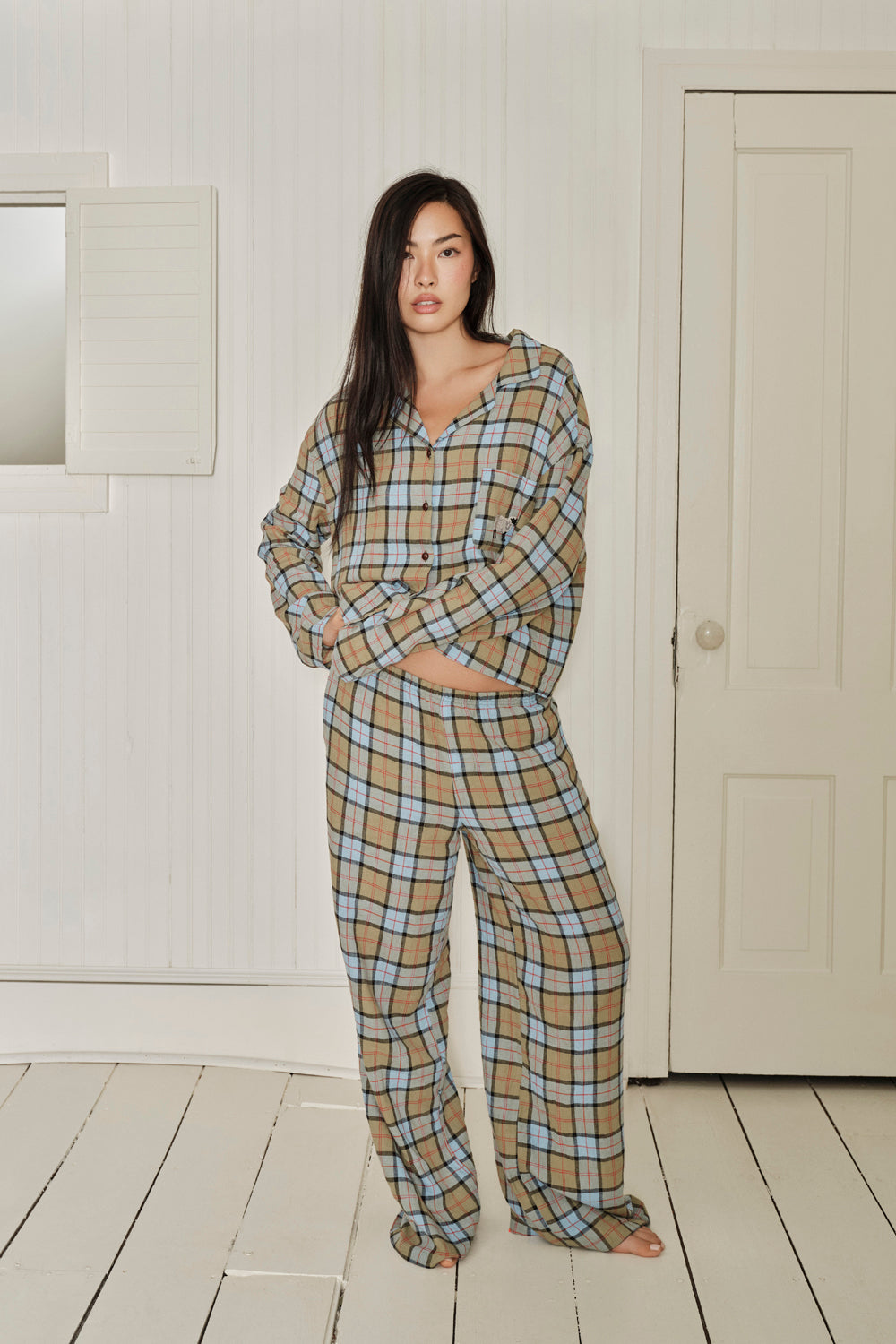 Woman wearing a plaid pajama set standing in a room with white walls and wooden floor. |M1
