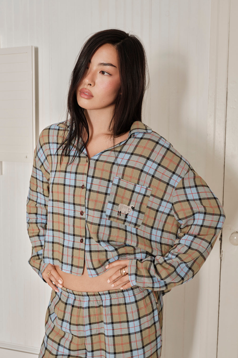 Woman wearing a plaid shirt and pants against a white wall. |M1