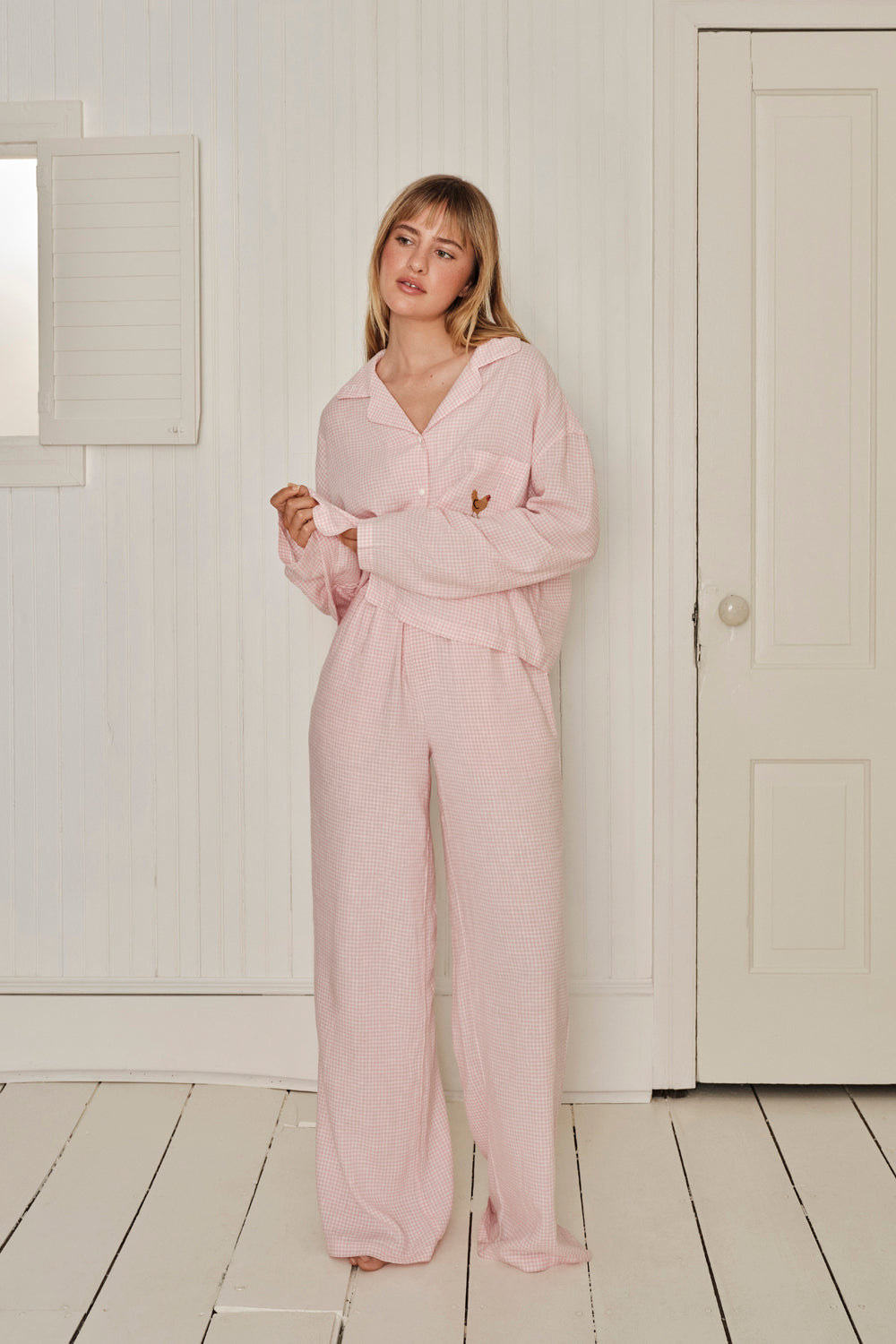 Woman wearing a pink gingham sleep set in a minimalistic room with white walls and wooden floor. |M1