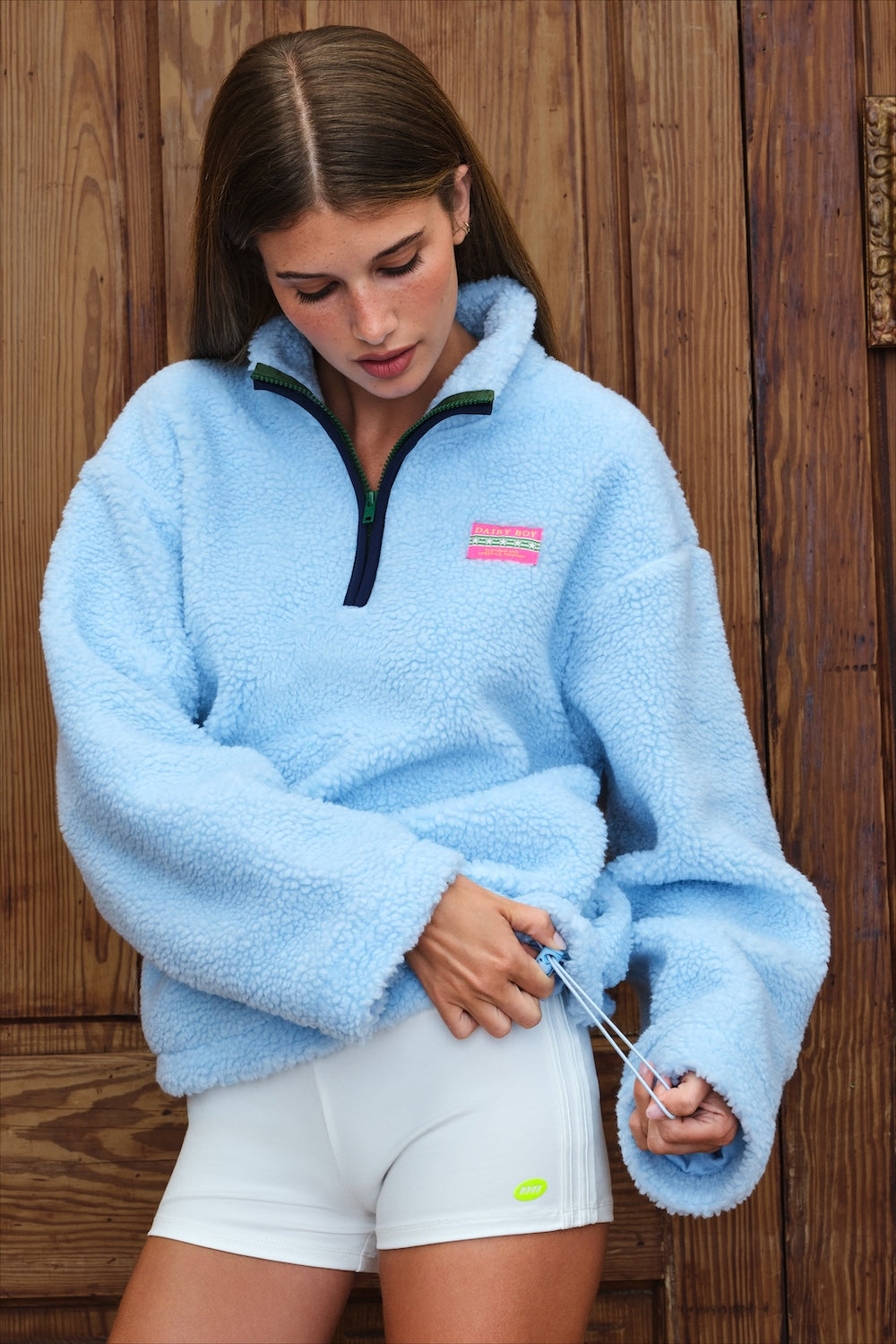 A woman wearing a light blue sherpa fleece quarter-zip pullover with a pink and green logo patch on the chest, paired with white athletic shorts, standing in front of a rustic wooden door.
|M1