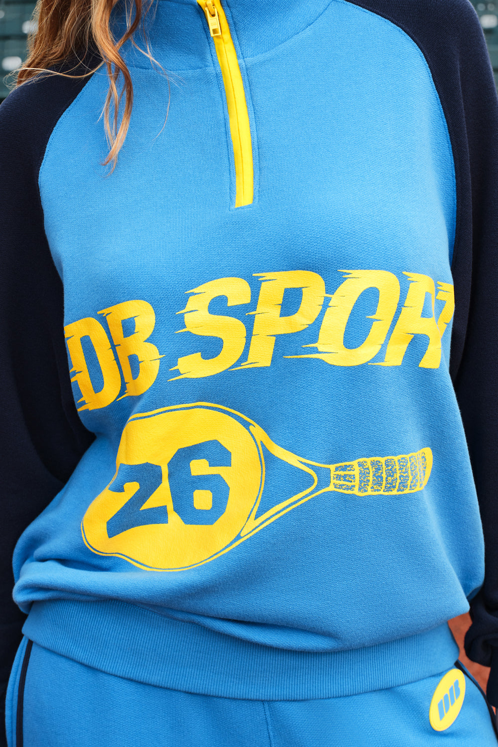 A close-up of a person wearing a bright blue and navy quarter-zip sweatshirt with a yellow graphic print that reads 'DB SPORT' and '26' inside a tennis racket outline.
|M1