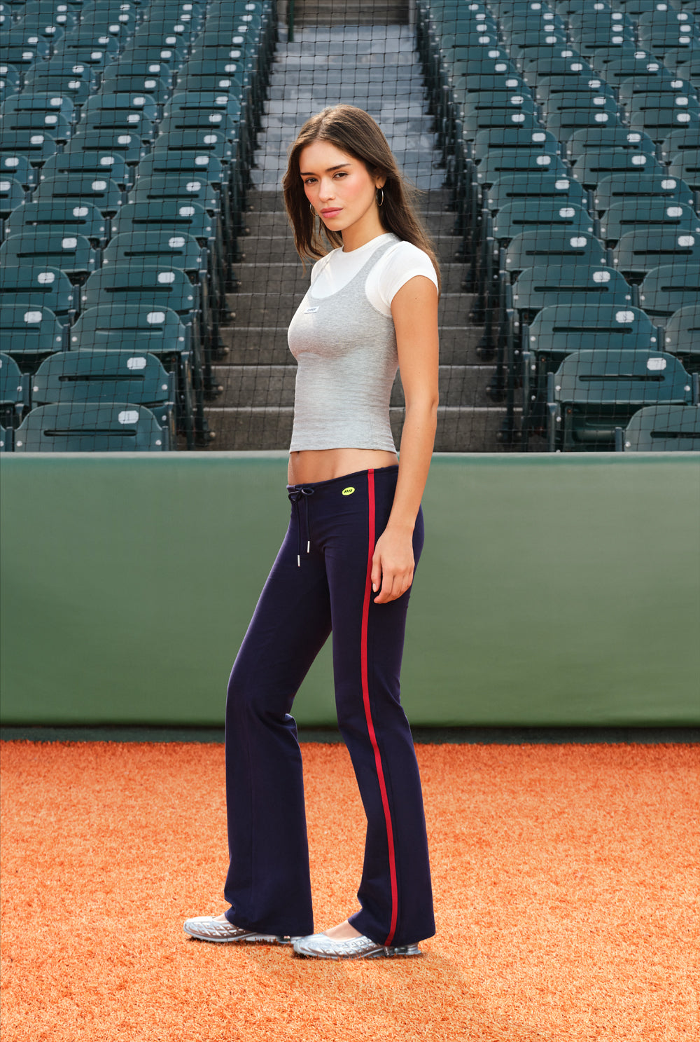 A woman stands on a baseball diamond wearing navy flared athletic pants with a red side stripe, a light gray short-sleeve crop top, and white sneakers. Empty green stadium seats rise in the background.
|M2