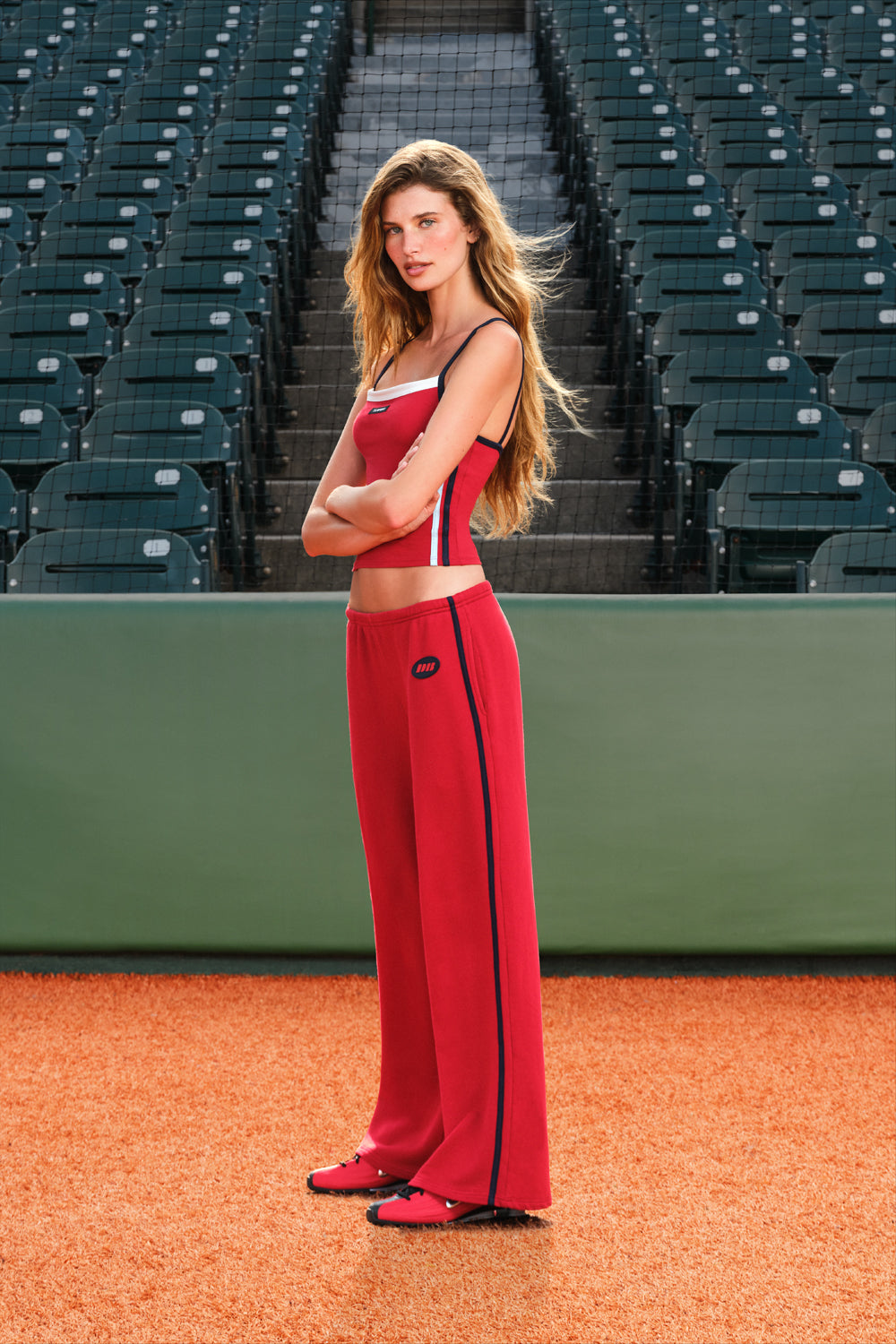 A woman stands on a baseball diamond wearing a matching red athletic set with white and black trim, consisting of a cropped tank top and wide-leg pants. Empty stadium seats are visible in the background.
|M1