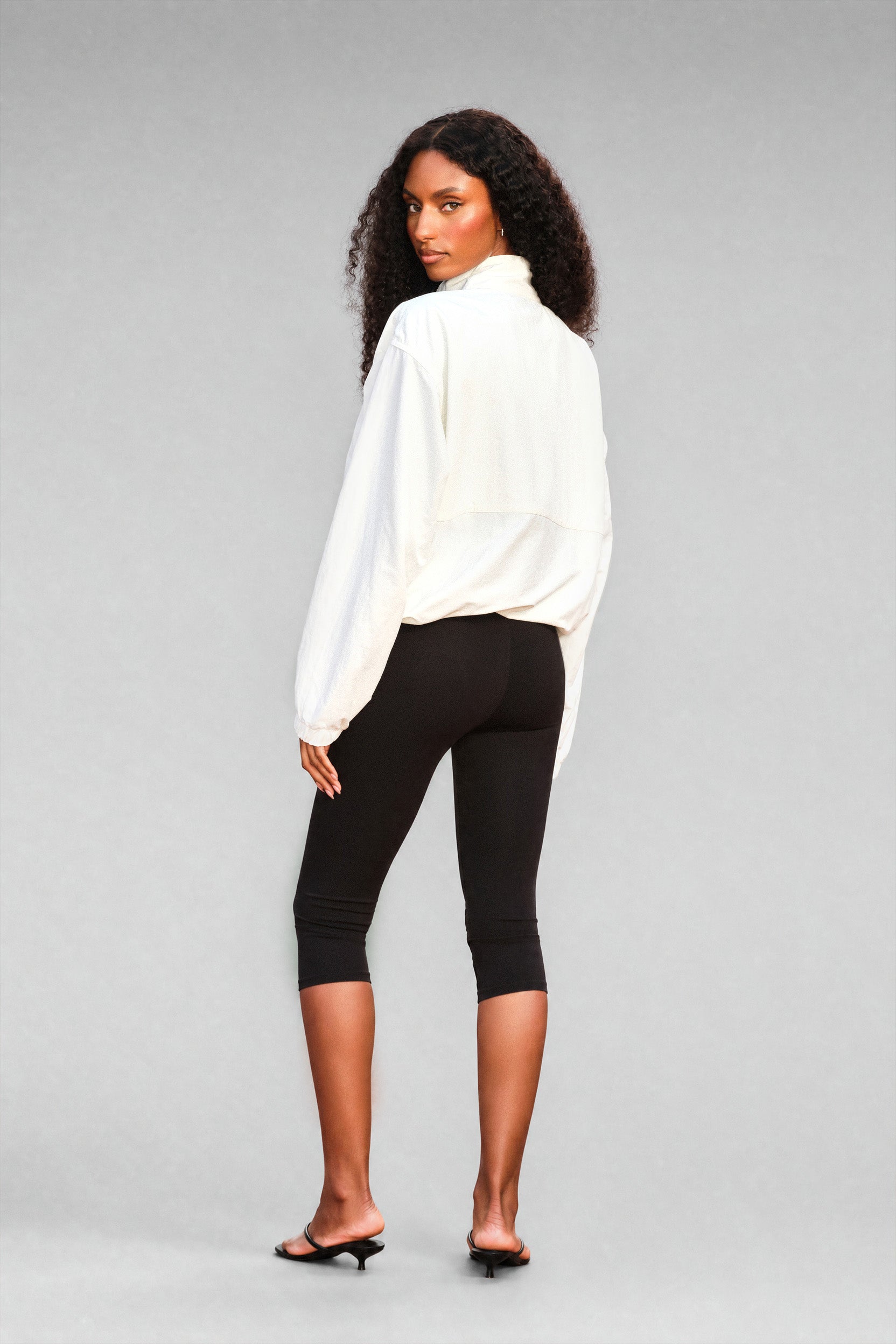 A woman with curly black hair is shown from the back wearing a white long-sleeve track jacket, black capri leggings, and black mule sandals against a solid gray studio background.
|M1