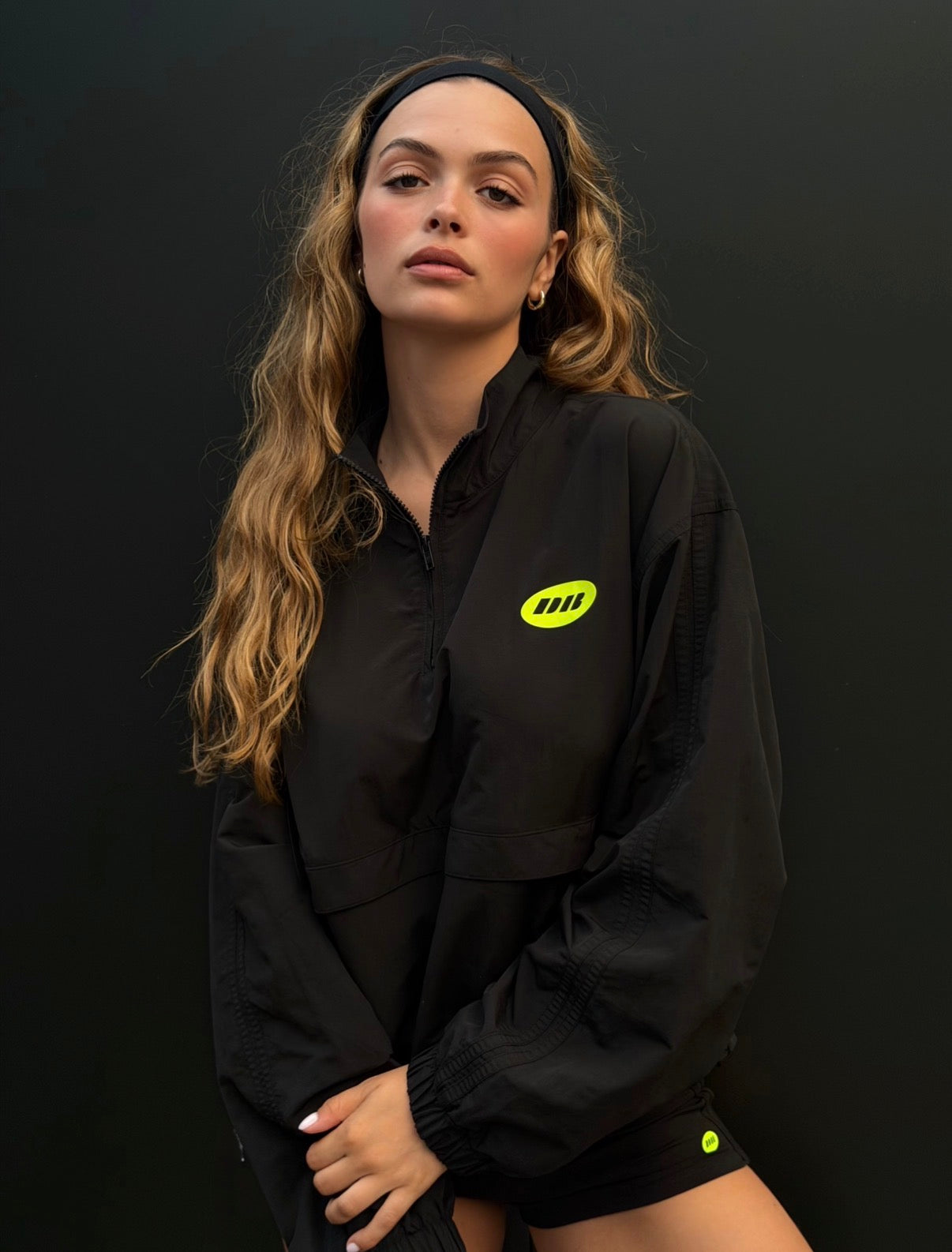 A woman modeling a black lightweight windbreaker jacket with a bright yellow-green logo on the left chest, paired with black shorts against a dark studio background.
|M1
