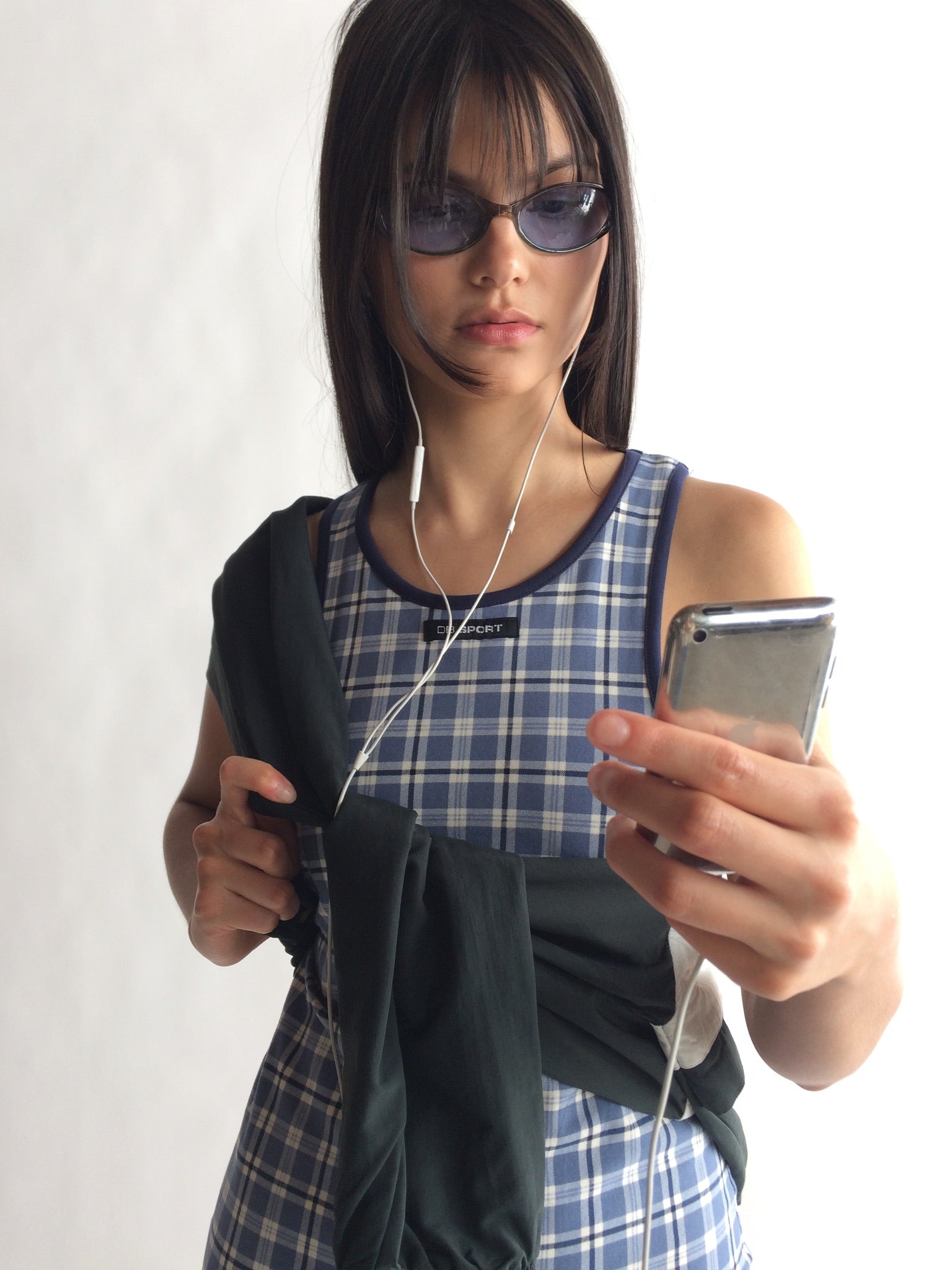 Woman wearing sunglasses, plaid dress, and holding a phone with earphones. |M2