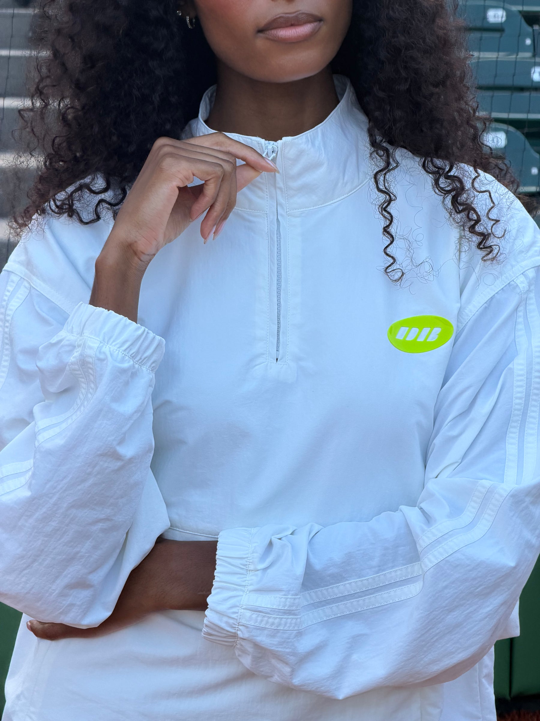 A woman wearing a white long-sleeve quarter-zip pullover jacket with a bright lime green oval chest logo featuring three diagonal stripes, posed against a dark green outdoor background.
|M1