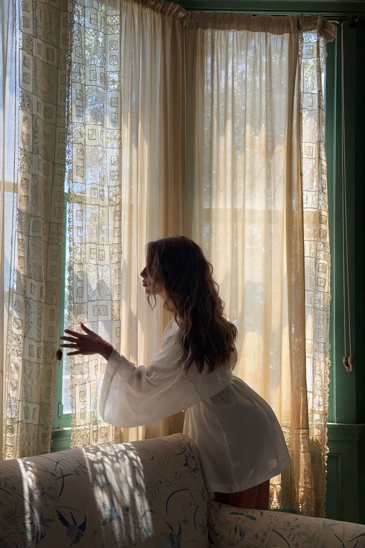 A woman in a white long-sleeve robe stands by a window with sheer cream curtains, looking out while wearing a white robe in a sunlit room.
|M1
