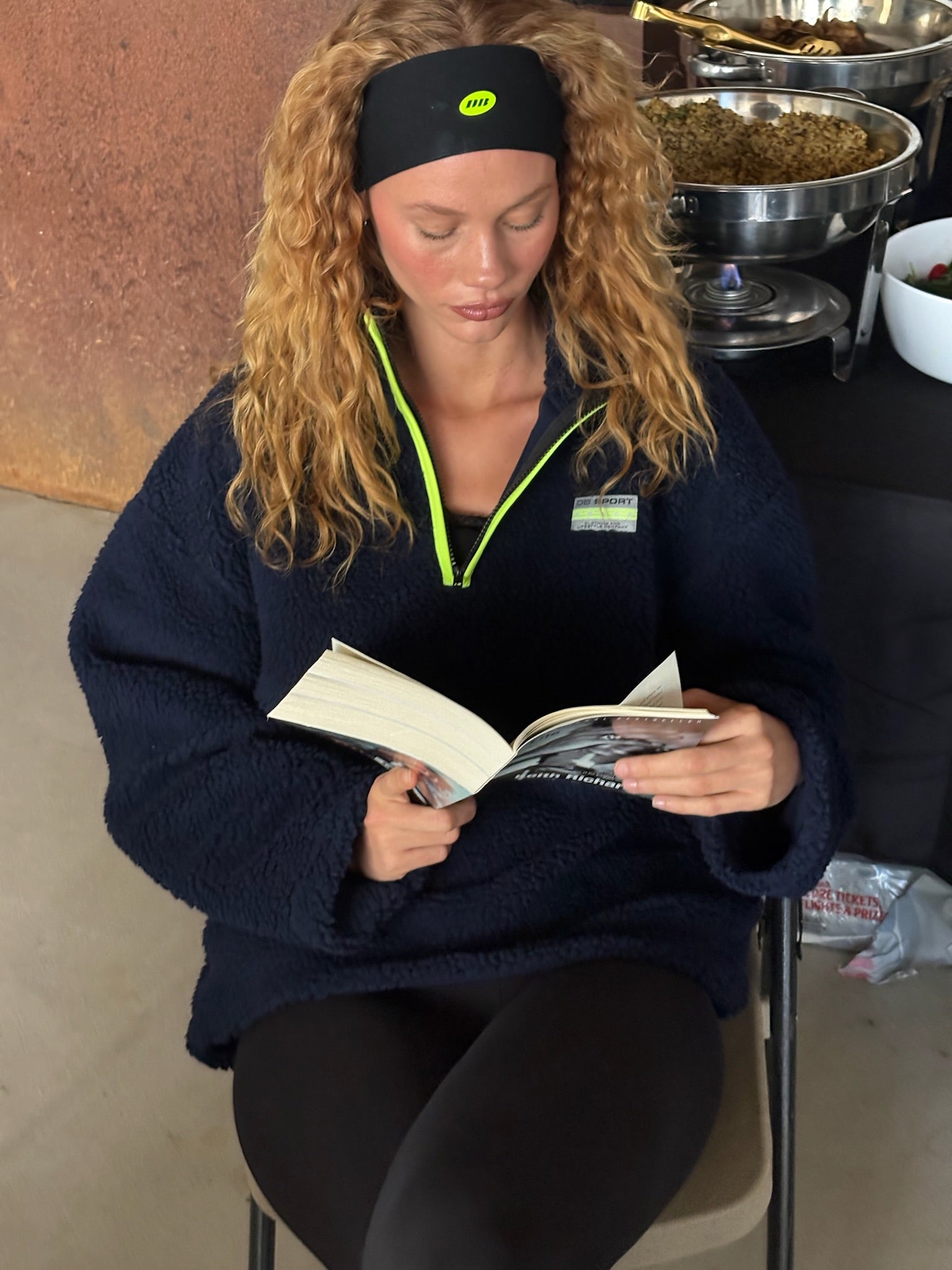 Woman reading a book in a casual setting with a headband and navy fleece. |M1