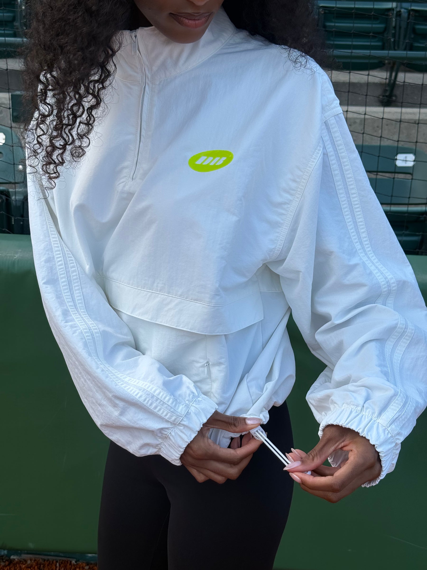 A woman wearing a white lightweight zip-up windbreaker jacket with a bright yellow-green oval chest logo, paired with black leggings, standing on an outdoor tennis court.
|M1